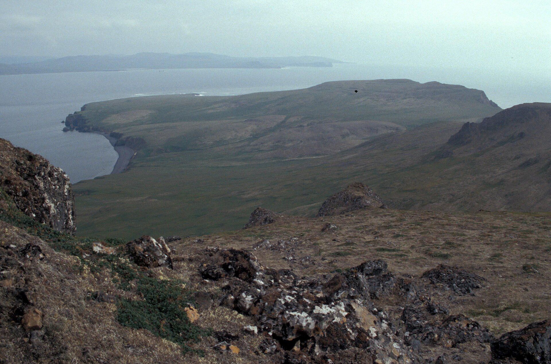 A bare green and brown island with cliffs dropping to the sea, and a second island faint in the mist behind it