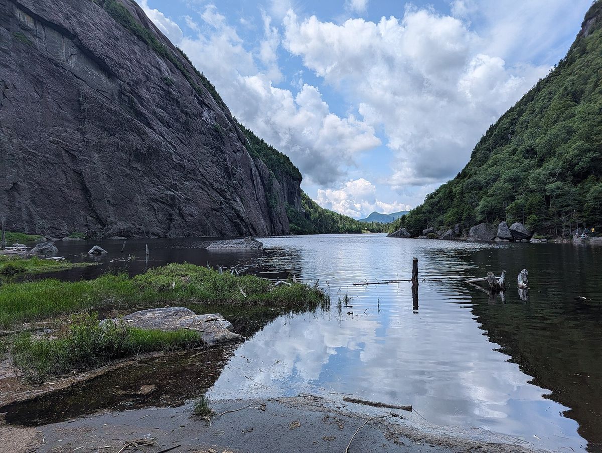 A long narrow mountain lake between a steep rock cliff and a forested slope in the Adirondacks, with clouds reflected in the water