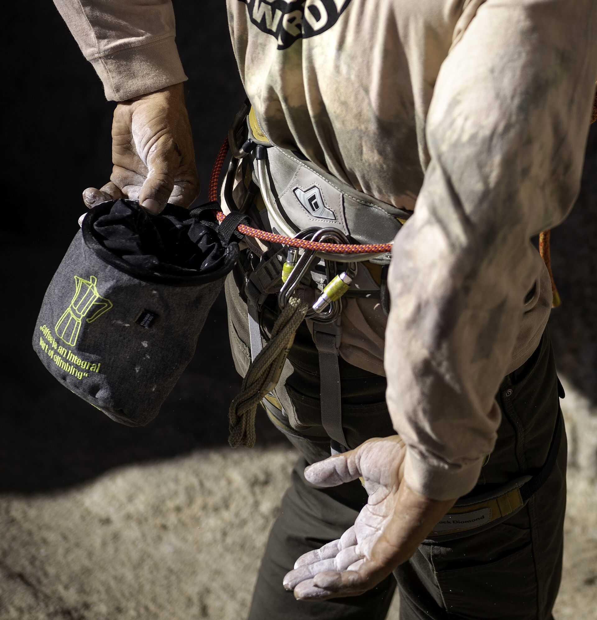 A rock climber seen from behind, dipping one hand into a bag of chalk. The other hand is open and white with chalk