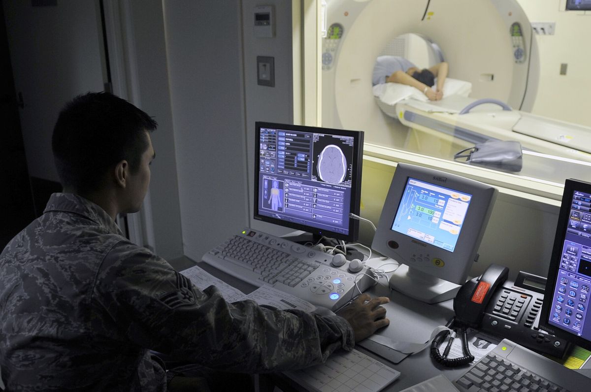 A technologist at a control desk, looking through a window at a person lying in a ring-shaped scanner