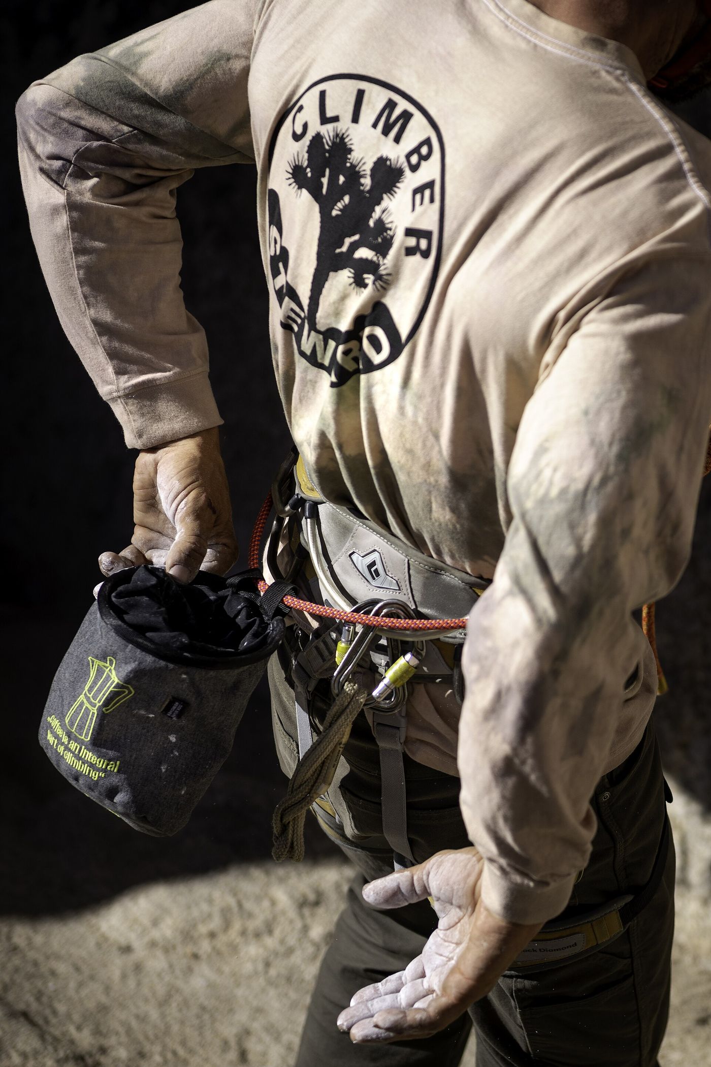 A rock climber dusting white chalk onto his open hands, with a chalk bag clipped to his belt