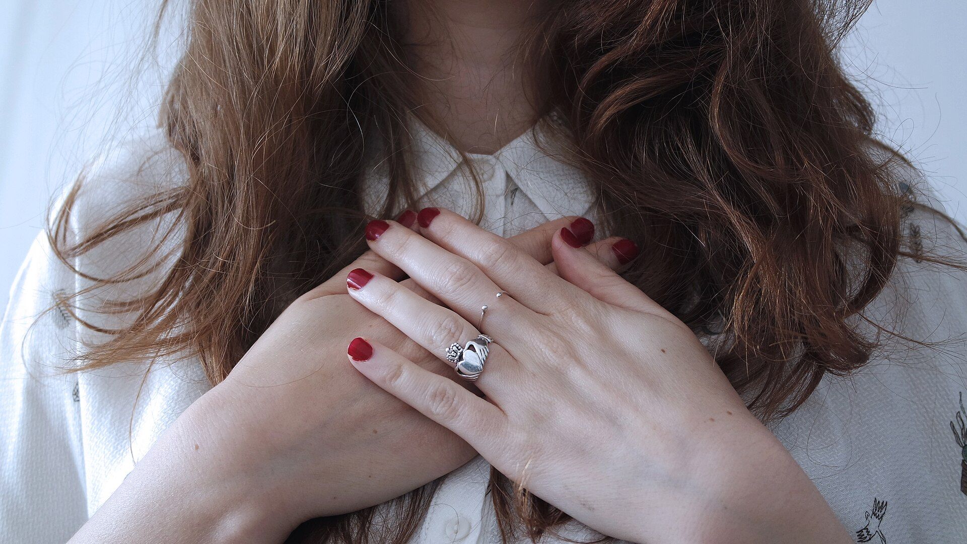 Two hands with red painted nails crossed over a white shirt, with long wavy brown hair falling around them