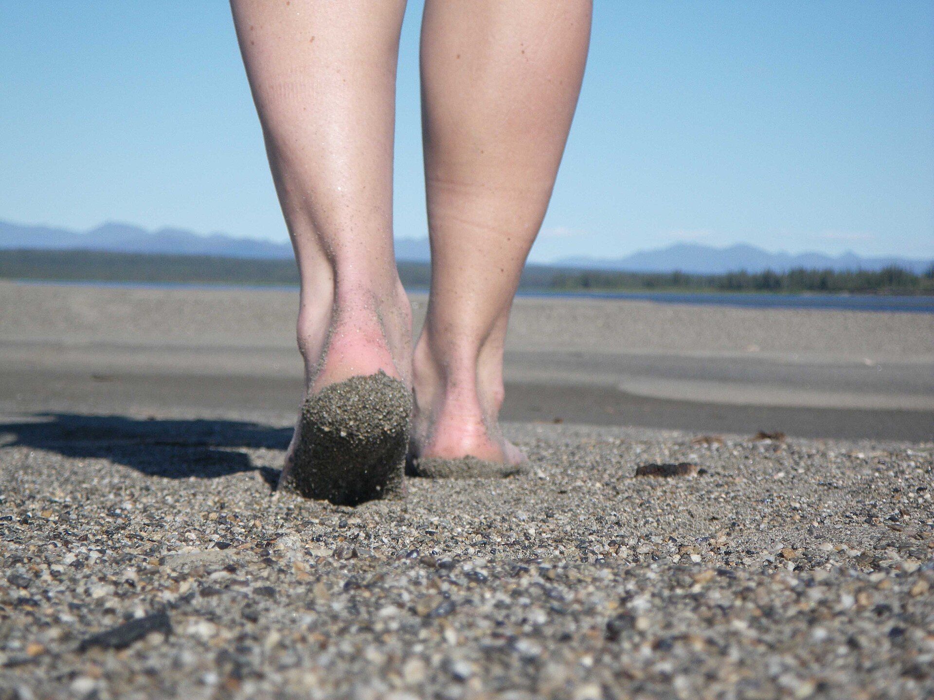 Two bare feet walking away across wet sand, one heel lifted and coated in sand