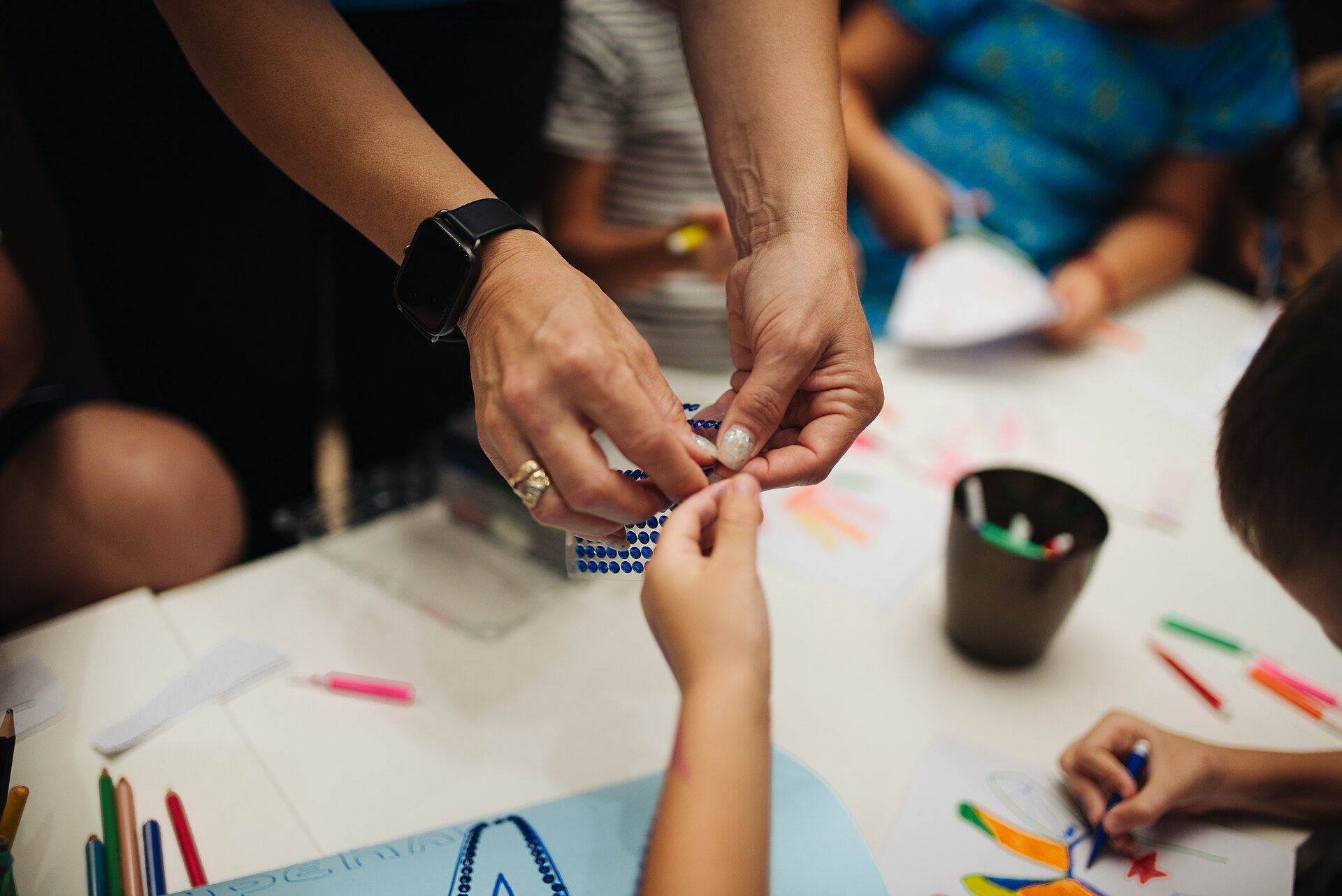An adult’s hands and a child’s hands working together on a paper craft at a table covered with markers