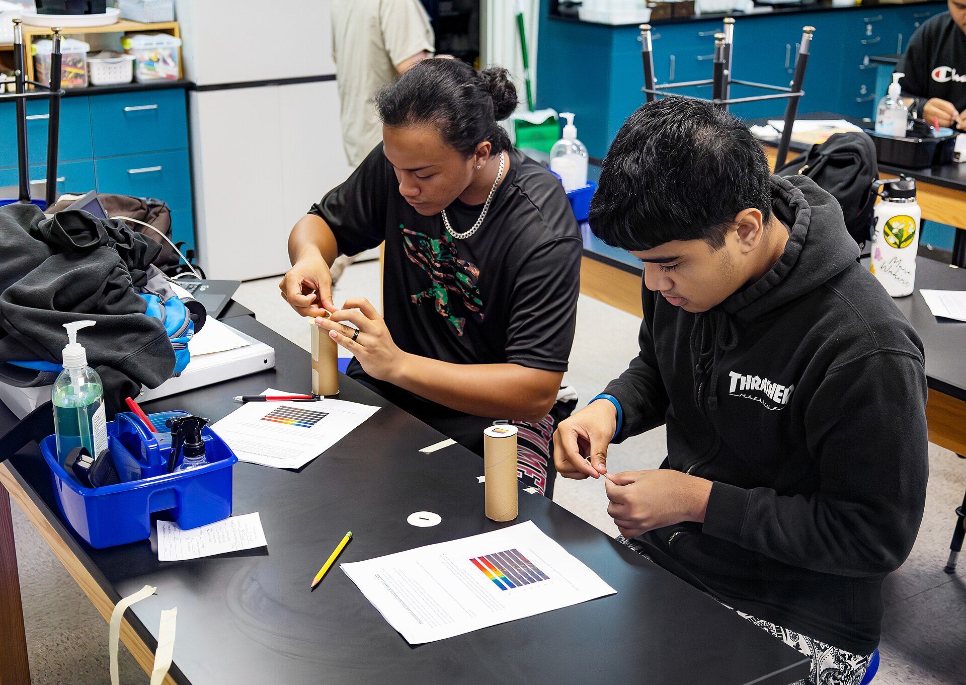 Two high school students at a lab table building spectroscopes from cardboard tubes, with a printed spectrum chart on the table